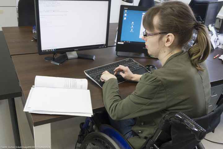 Disabled worker in a wheelchair typing at keyboard while looking at two computer screens.