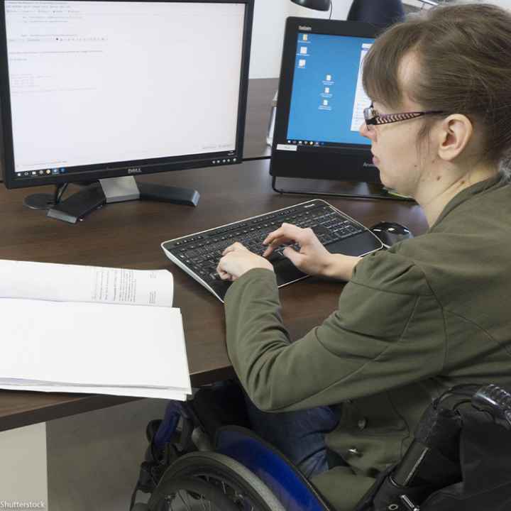 Disabled worker in a wheelchair typing at keyboard while looking at two computer screens.